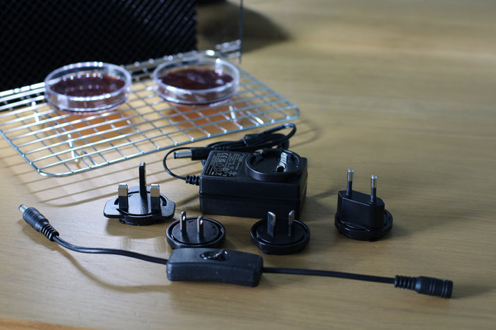 A small laminar flow hood power adapter with multiple international plug attachments, placed on a wooden surface. A stainless steel rack and Petri dishes are visible in the background.
